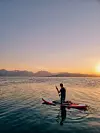 En person står på et padlebrett på stille sjø i solnedgang foran fjell.